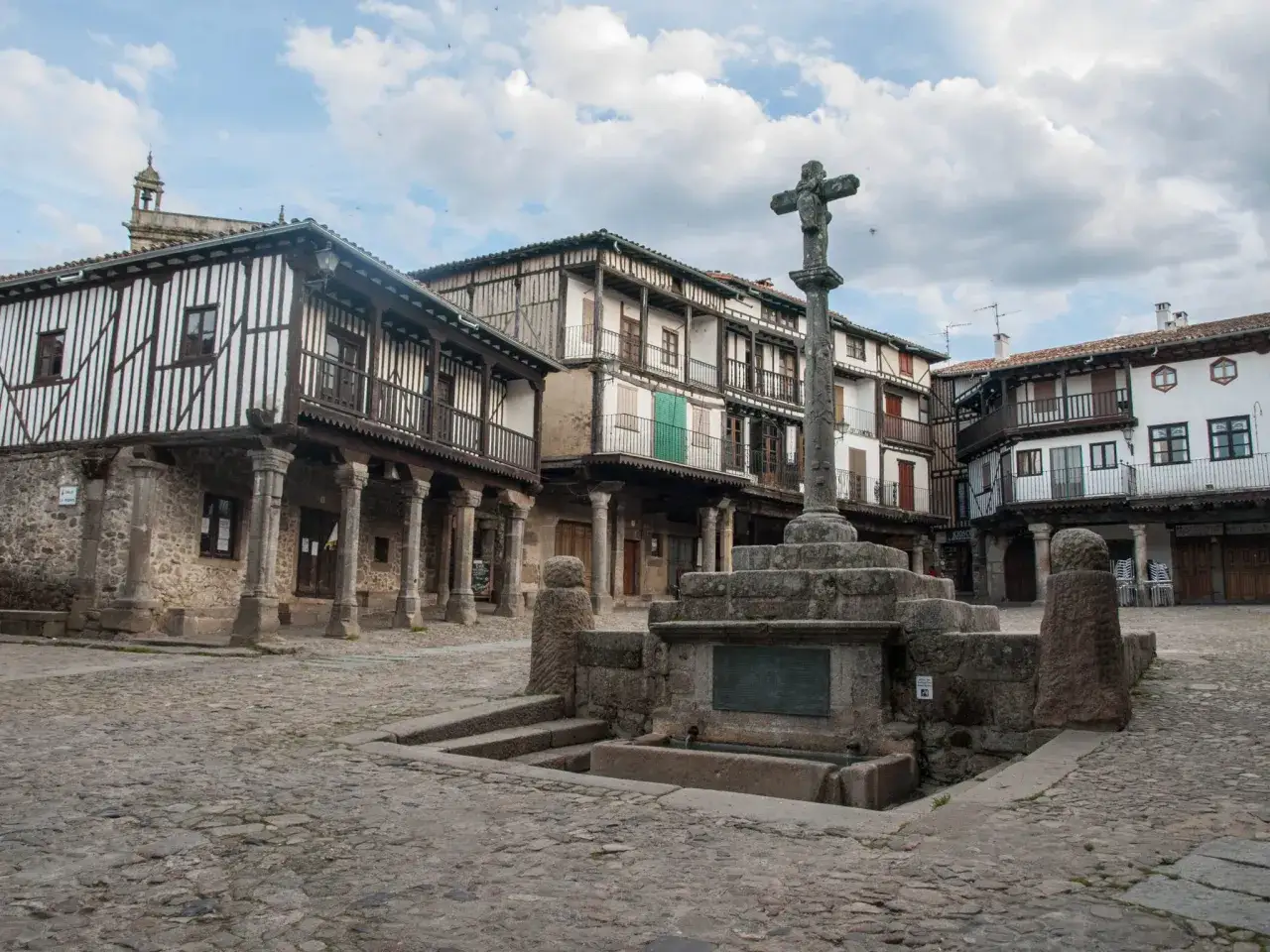 Plaza con fuente y cruz de piedra, rodeada de casas con balcones de madera, típicas de los pueblos de la sierra de Francia.