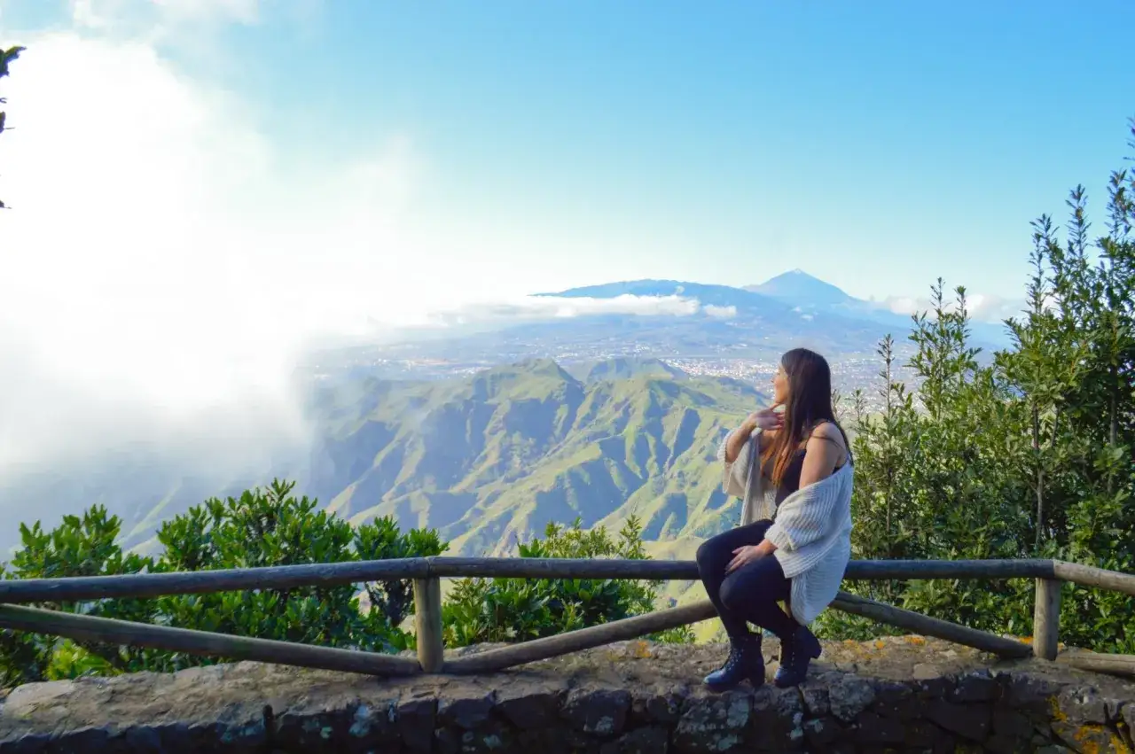 Mujer contempla el monte del agua, un paisaje verde y montañoso bajo un cielo azul.