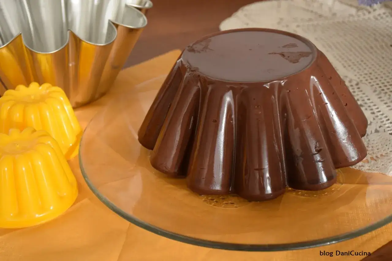 Budino al cioccolato fatto in casa, lucido e invitante, pronto per essere gustato. Accanto, due piccoli budini gialli e uno stampo metallico.