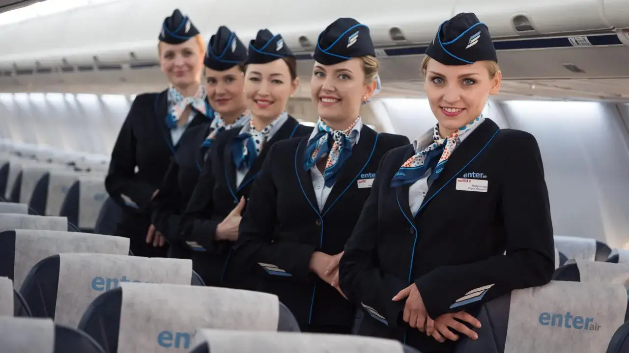 Pięć uśmiechniętych stewardess w granatowych mundurach i czapkach, gotowych do lotu. Marzysz, jak zostać stewardessą? To może być Twój pierwszy krok!