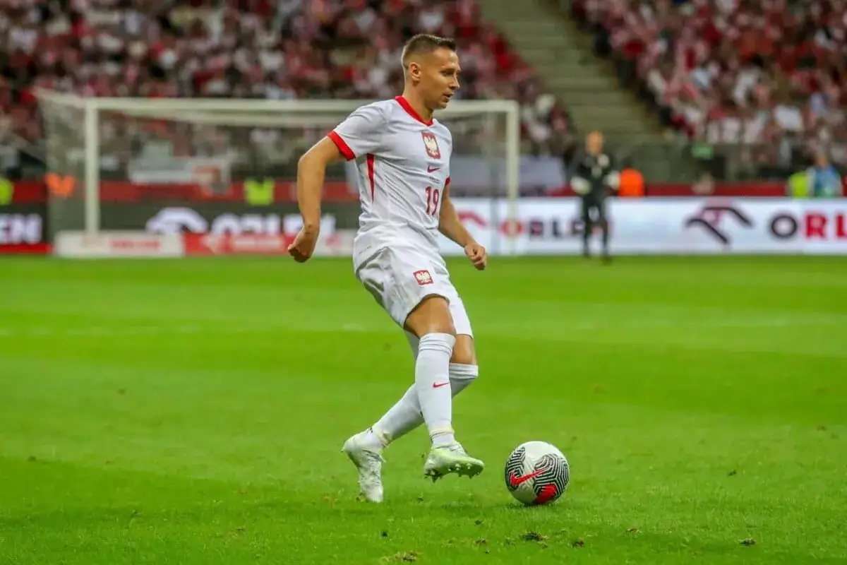 Frankowski piłkarz w białej koszulce z numerem 19 drybluje na zielonej murawie stadionu, gotowy do strzału.