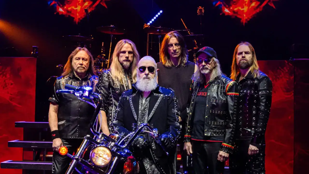 Zespół Judas Priest na scenie, gotowy do koncertu. Wszyscy w skórzanych kurtkach, jeden z nich siedzi na motocyklu.