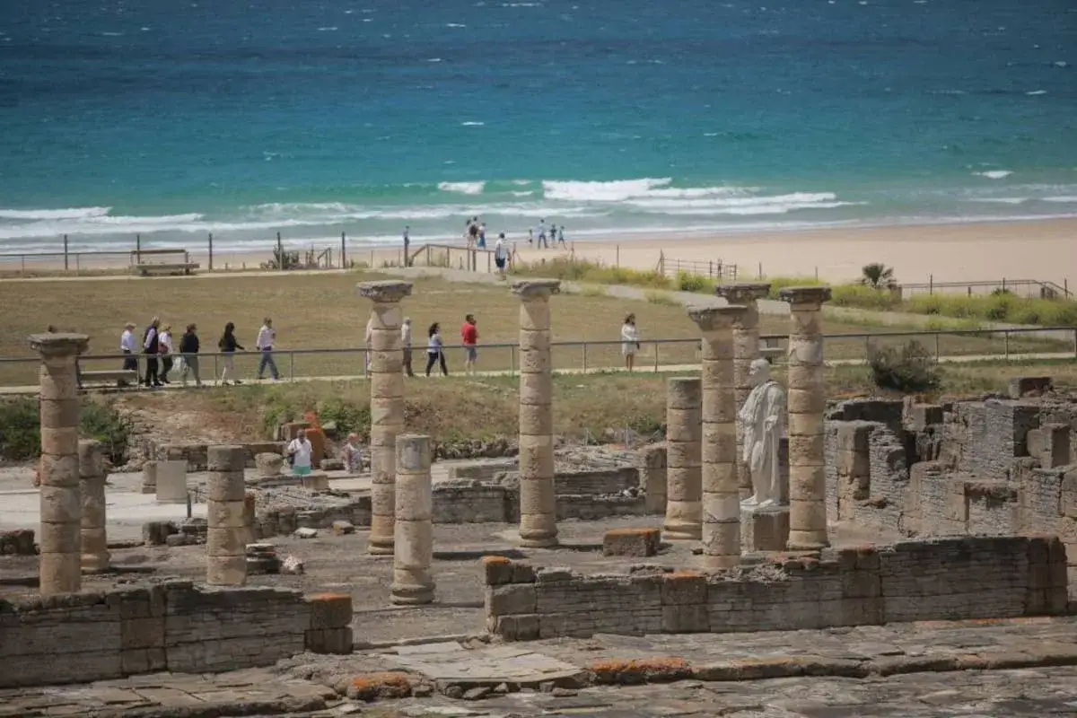Columnas y muros de ruinas romanas en Cádiz, con el mar azul y la playa al fondo. Turistas pasean entre los vestigios.