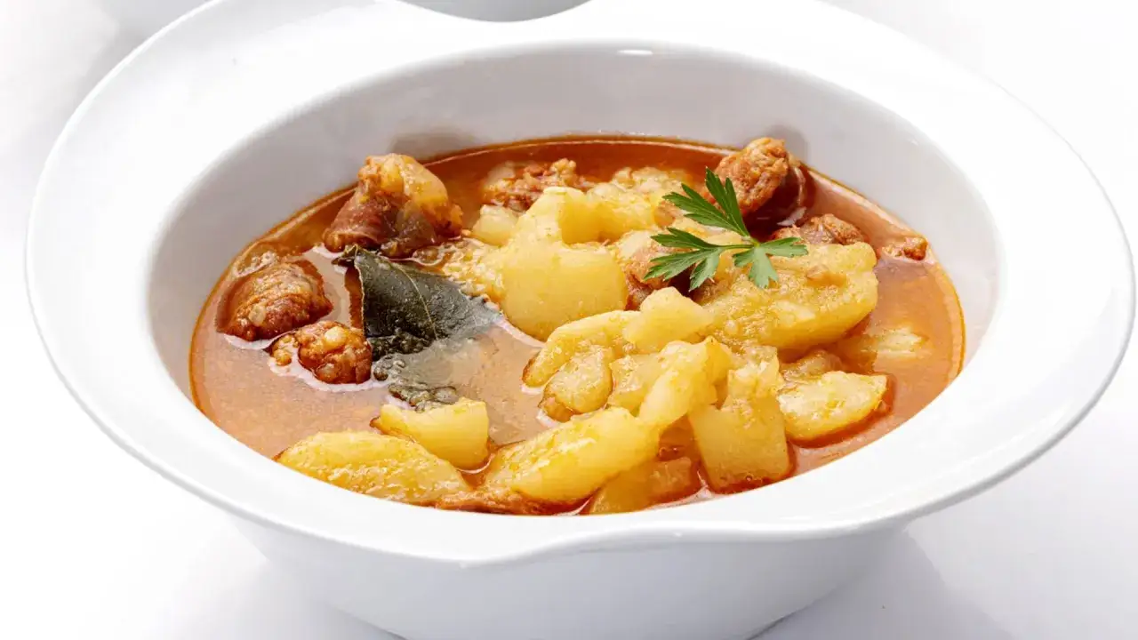 Un tazón blanco lleno de patatas a la riojana, con trozos de chorizo y carne en un caldo rojizo.