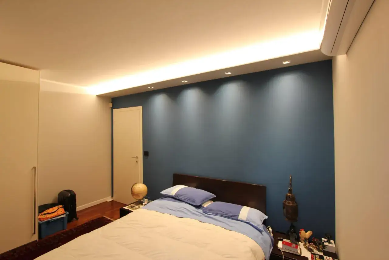 Camera mostra una camera da letto con un letto, un armadio e una parete blu. La testata letto in cartongesso è illuminata da faretti.