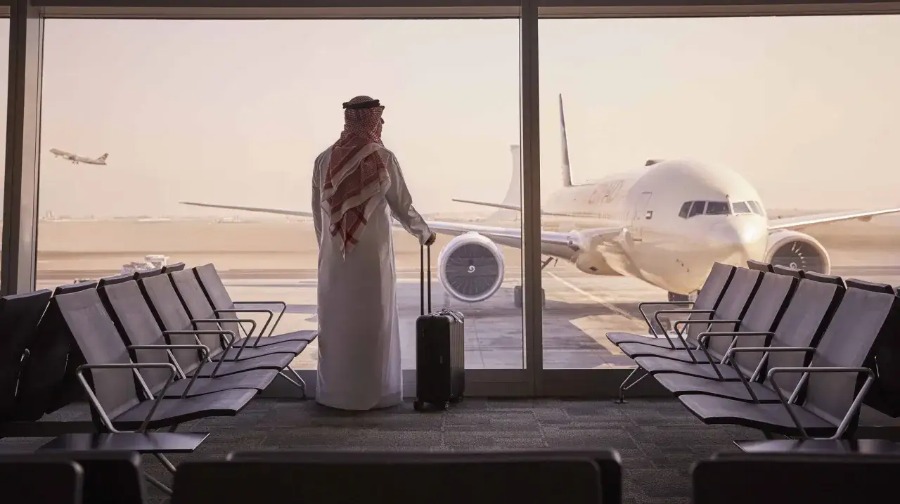 Mężczyzna w tradycyjnym stroju z walizką czeka na lotnisku AUH Zayed International. Samolot Etihad czeka na płycie.