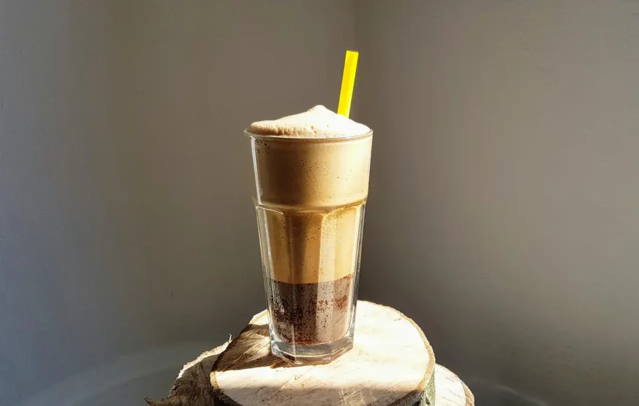 Pyszny frappe, czyli mrożona kawa z pianką, w szklance ze słomką, na tle ściany.