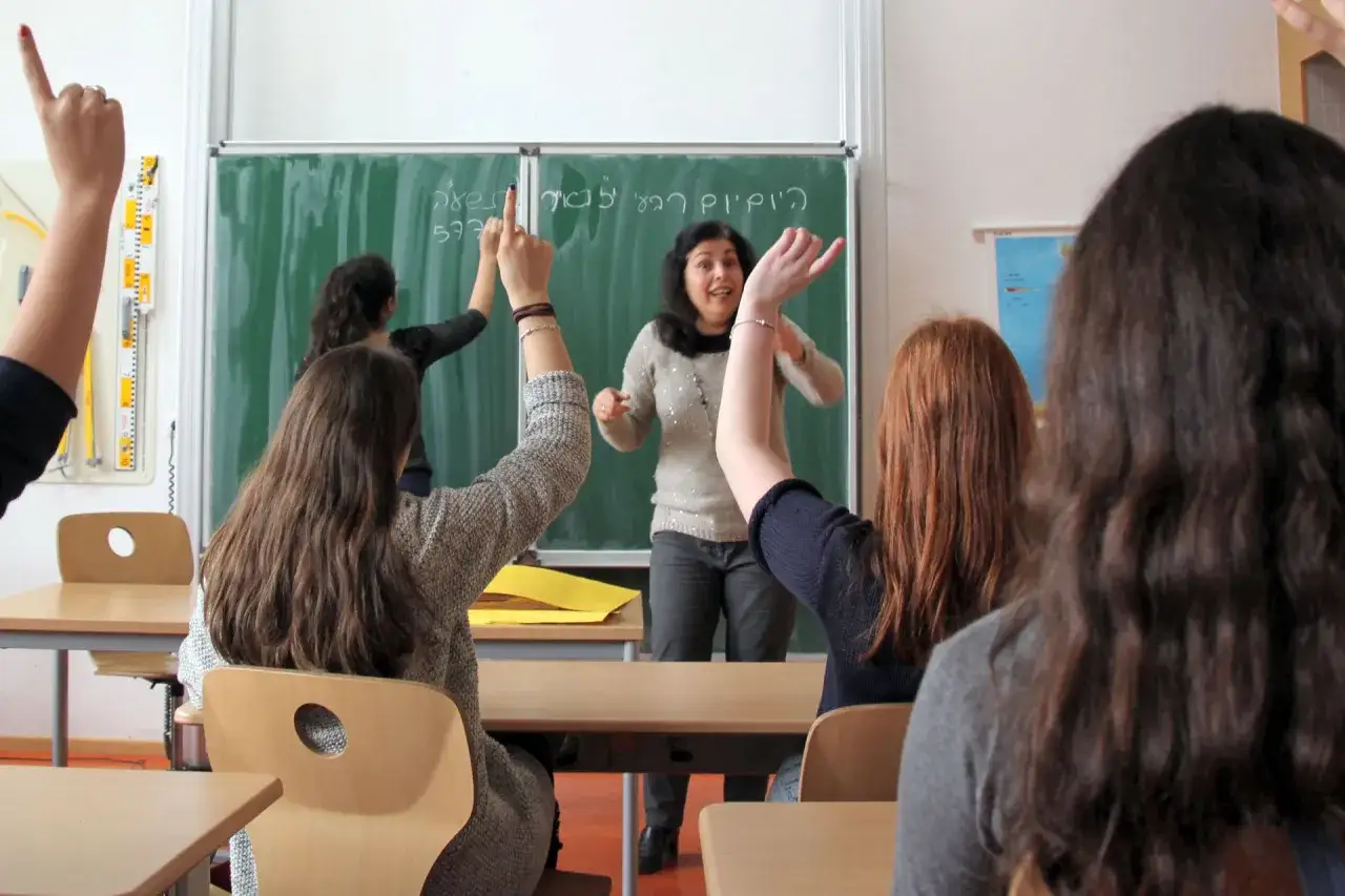 Schülerinnen heben die Hände, um Fragen zu beantworten. Die Lehrerin erklärt etwas auf Hebräisch, was das **fremdsprachen lernen** in dieser Klasse zeigt.