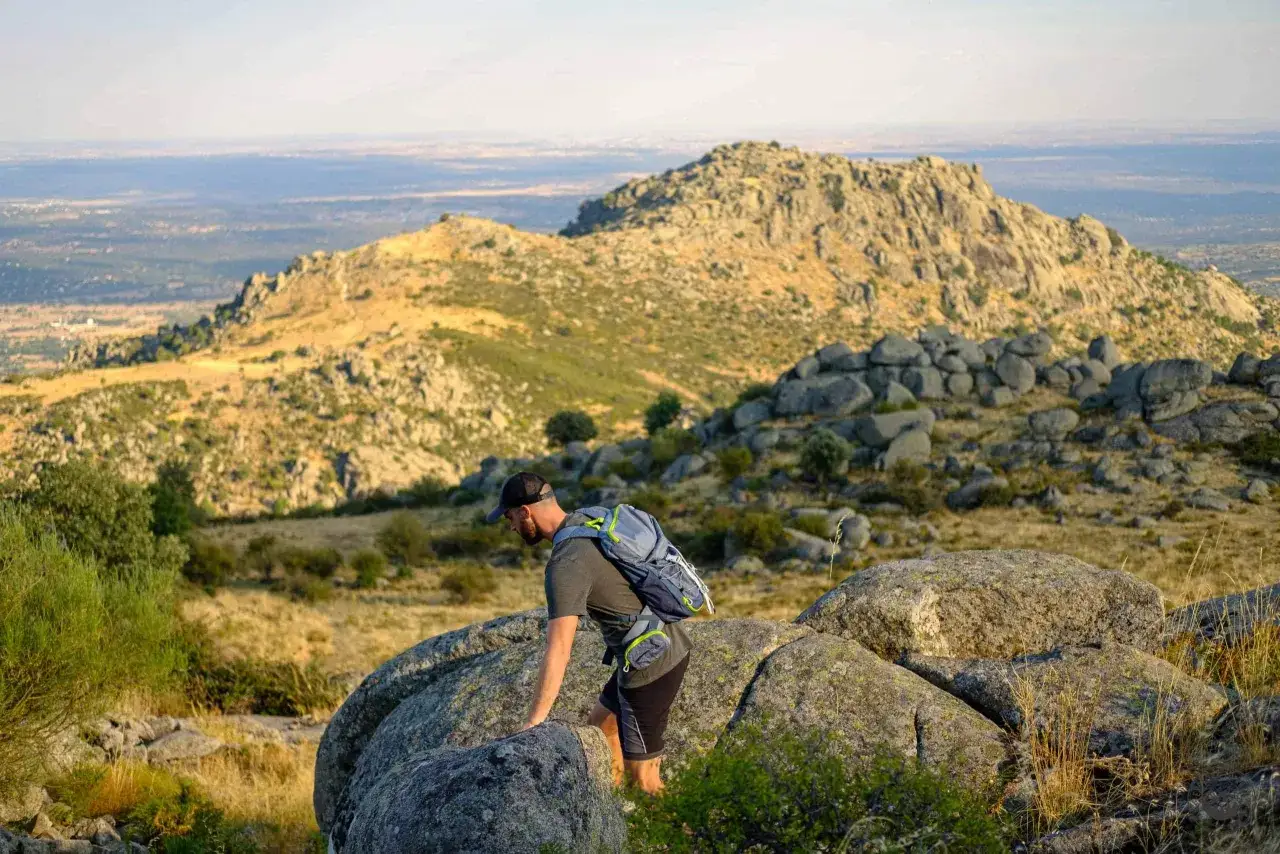 Un excursionista con mochila asciende por un terreno rocoso en la ruta Las Machotas, con un paisaje monta&ntilde;oso y un valle al fondo.