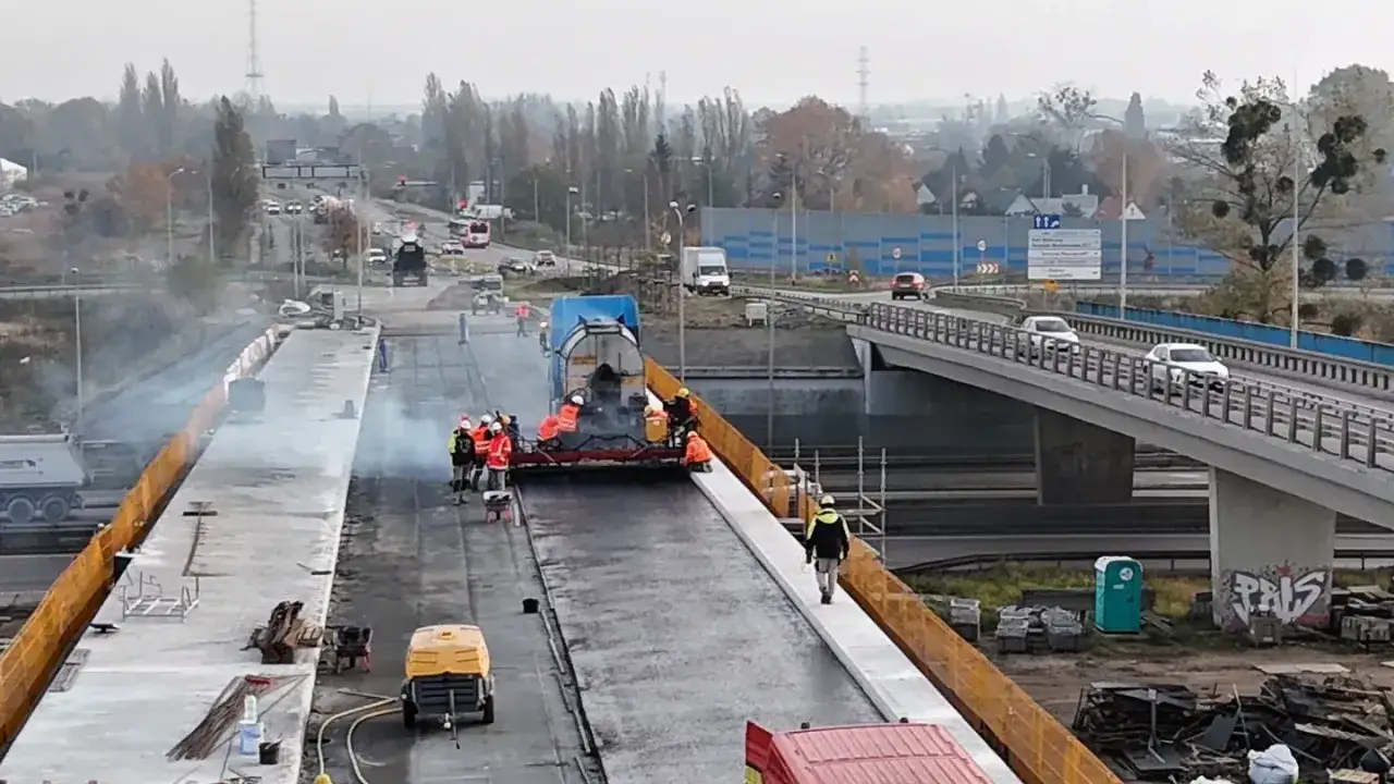 Budowa estakady w toku. Pracownicy układają nawierzchnię drogi, a w tle widać ruchliwy most.