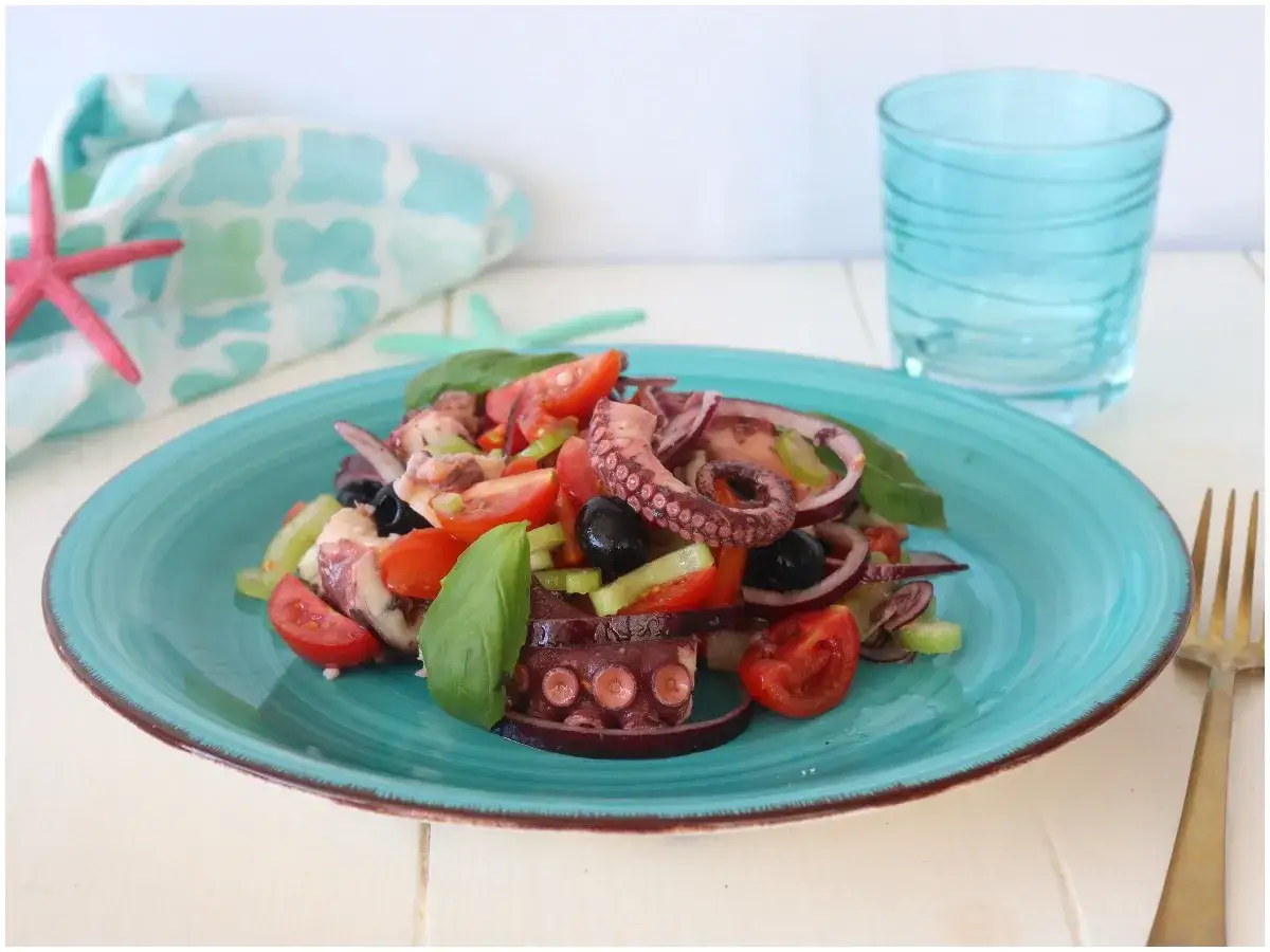 Delizioso polpo alla catalana su piatto turchese, con pomodorini, olive nere, cipolla rossa e basilico. Fresco e invitante.