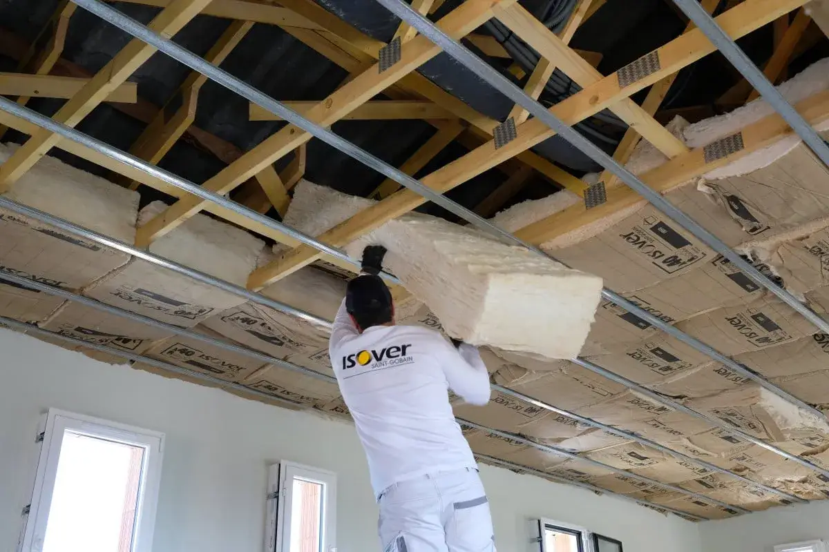 Un ouvrier installe un panneau de fibre de verre pour isoler un plafond. La structure en bois et les rails m&eacute;talliques sont visibles.