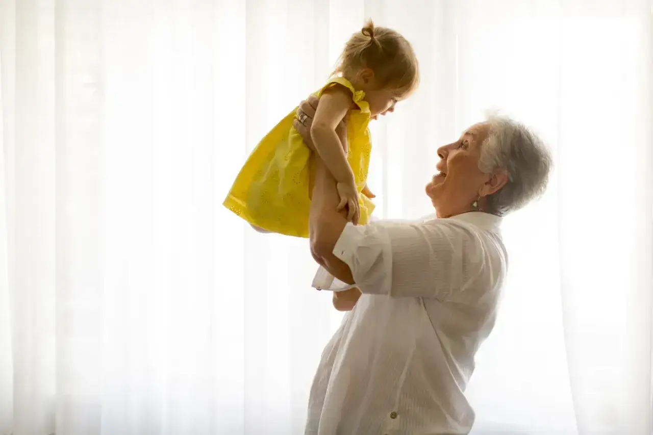 Uśmiechnięta babcia podrzuca w górę wnuczkę w żółtej sukience. Radość i miłość w tych babciowych chwilach.
