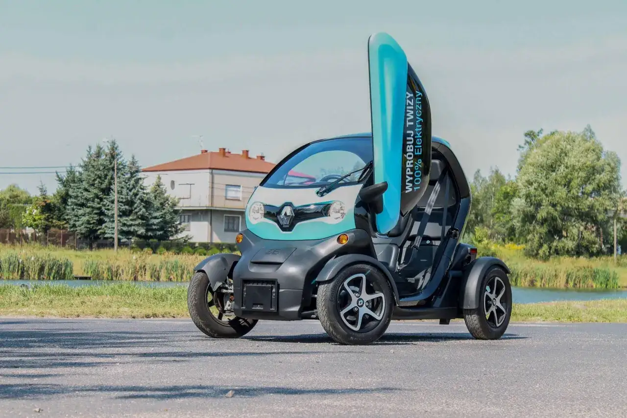 Mały, elektryczny Renault Twizy z otwartymi drzwiami, gotowy do jazdy nad jeziorem.