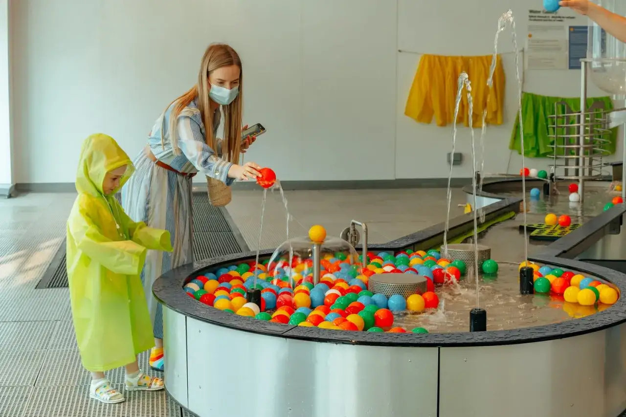 Wesoła zabawa w muzeum dla dzieci. Mama w maseczce i dziecko w żółtym płaszczu wrzucają kolorowe piłeczki do wodnej instalacji.