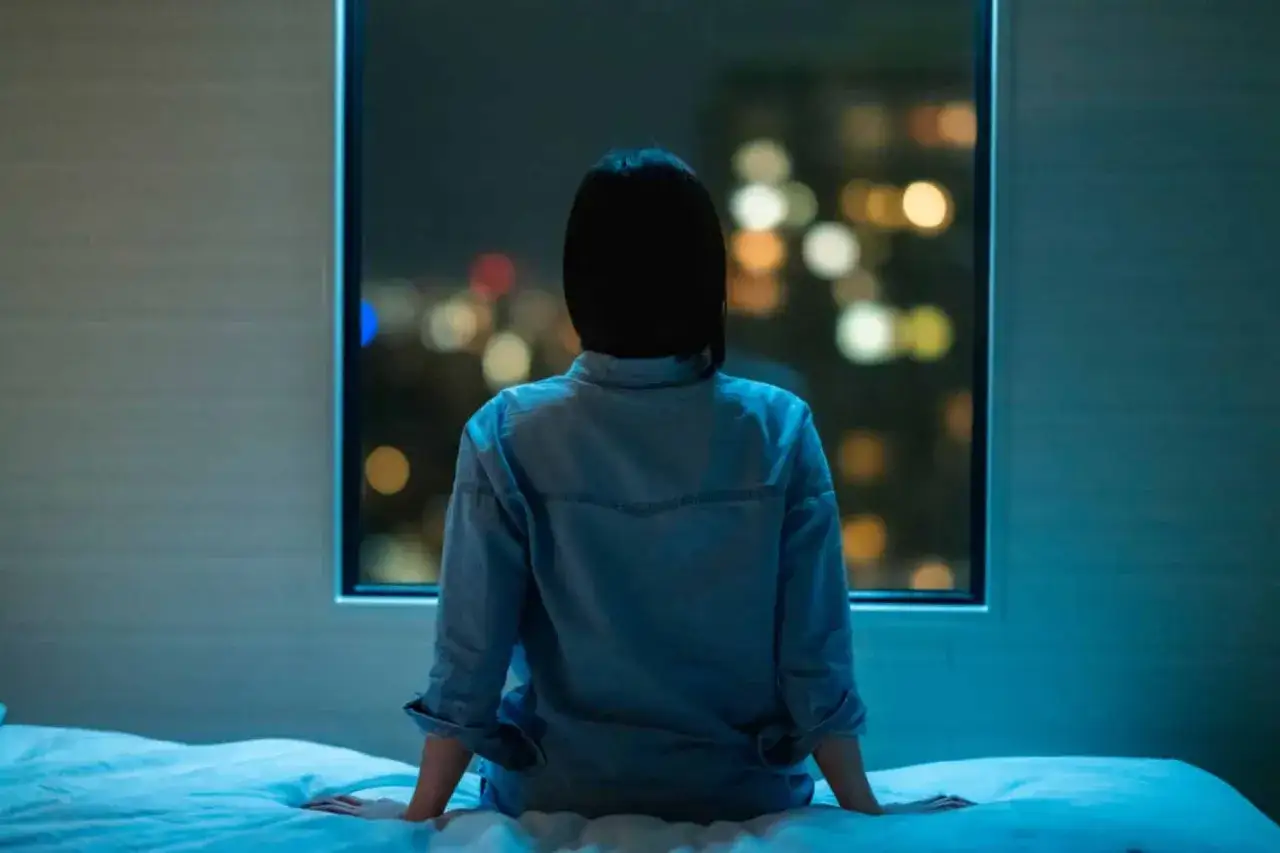 Kobieta siedzi na łóżku, patrząc na nocne miasto. Kolejna noc pełna bezsenności.