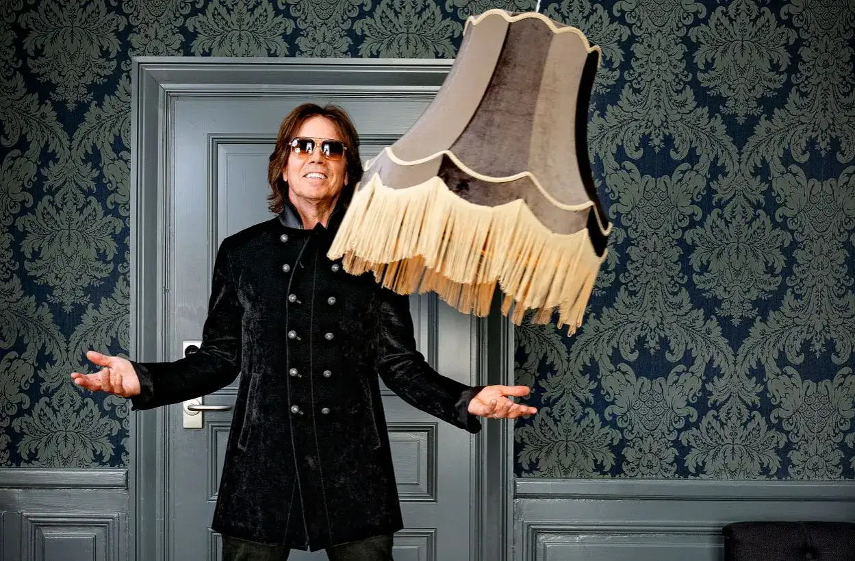 Joey Tempest, med sitt karakt&auml;ristiska leende och solglas&ouml;gon, poserar framf&ouml;r en d&ouml;rr med en lampa som sv&auml;var i luften.