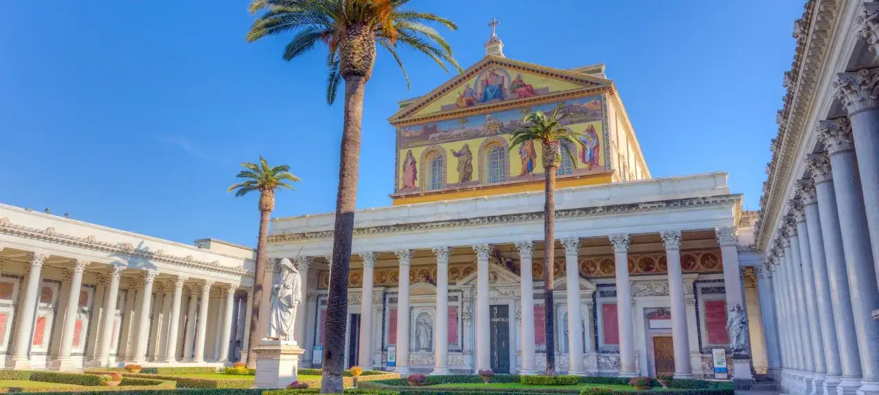 La Basilica di San Paolo Fuori le Mura, con le sue maestose colonne e palme, sotto un cielo azzurro.