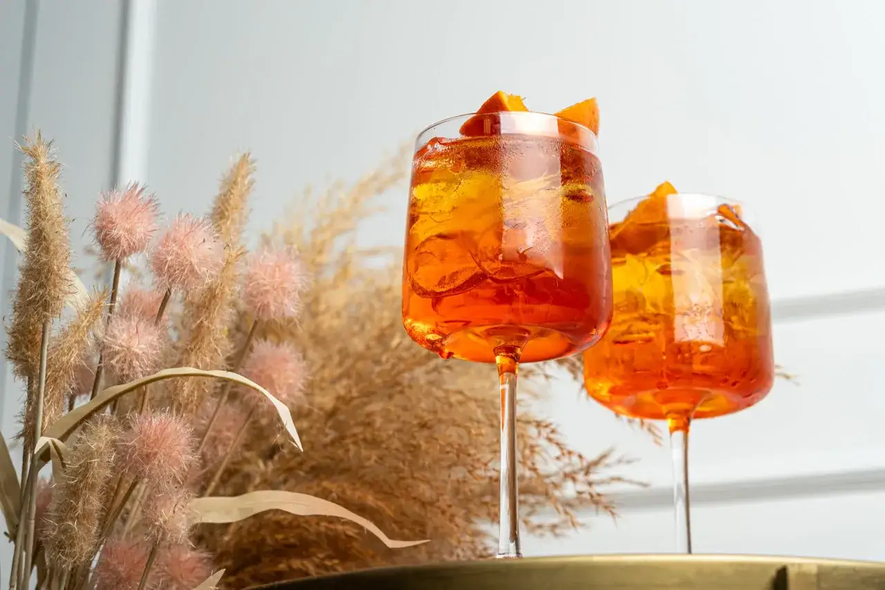 Tv&aring; glas med Aperol Spritz, fyllda med is och apelsinskivor, st&aring;r framf&ouml;r torkade gr&auml;sstr&aring;n. Vilket sodavatten till Aperol Spritz?