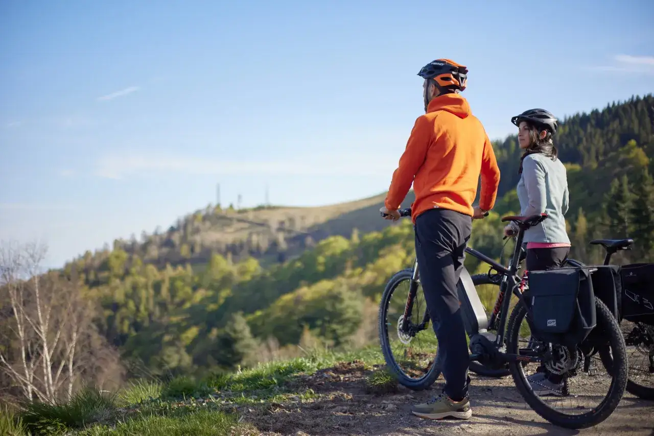 Coppia in bici sul cammino di Oropa, con vista panoramica su colline boscose e cielo azzurro.