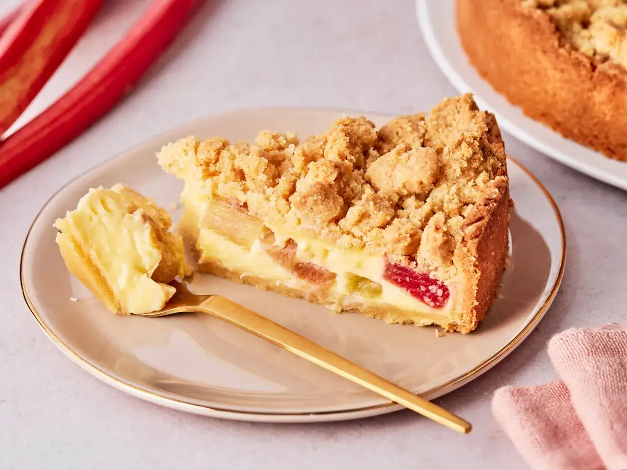 Ein saftiges St&uuml;ck Rhabarberkuchen mit Pudding und Streuseln auf einem Teller, daneben ein goldener L&ouml;ffel und frische Rhabarberstangen.