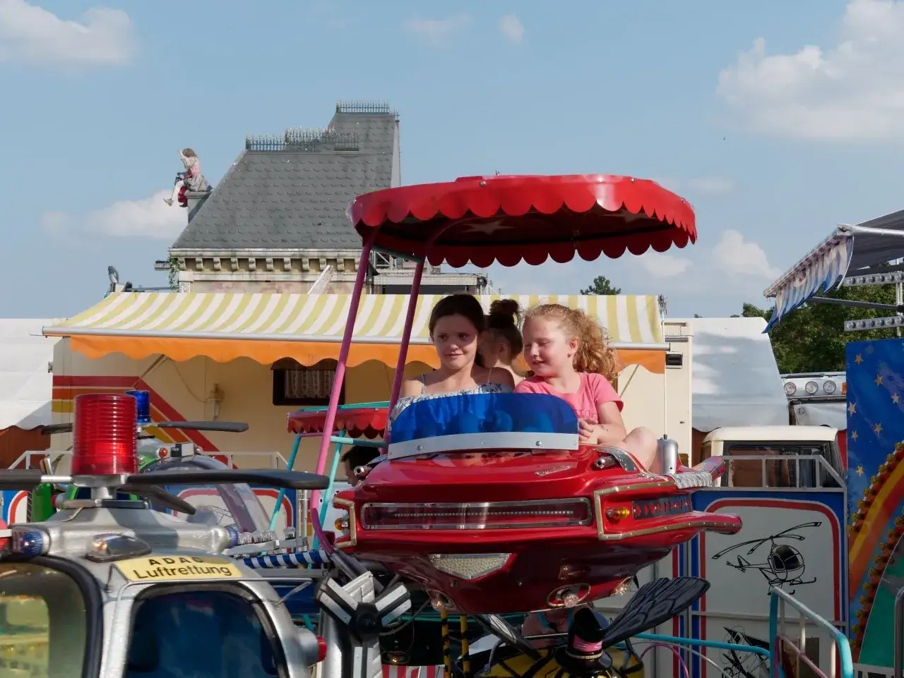 Zwei Mädchen auf einem Fahrgeschäft in Regensburg mit Kindern. Ein rotes Flugzeug mit Propellern und Blaulicht.