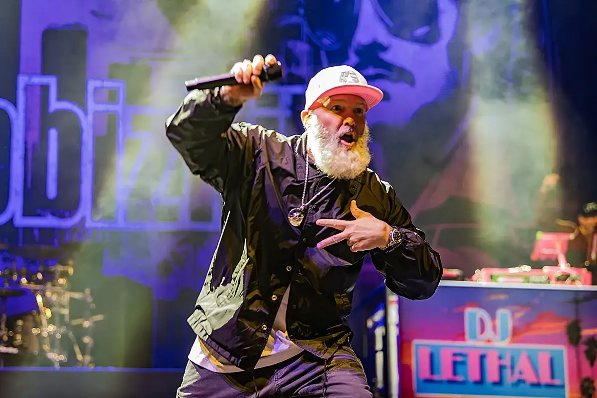 Energetyczny wokalista z brodą, w czarnej kurtce i różowej czapce, śpiewa na scenie podczas jednego z koncertów Limp Bizkit.