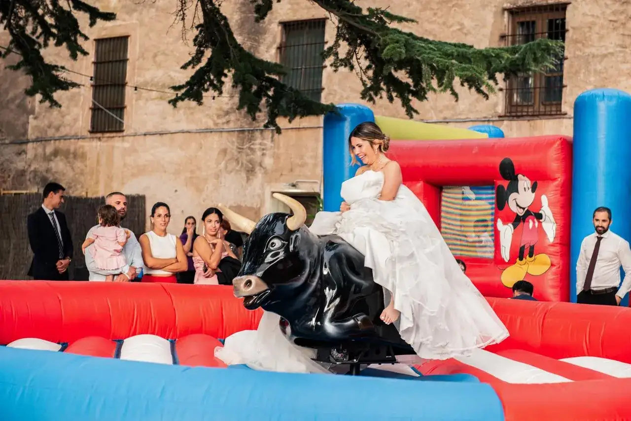 Novia sonriente en un toro mecánico, rodeada de invitados. ¡Ideas para bodas divertidas y memorables!