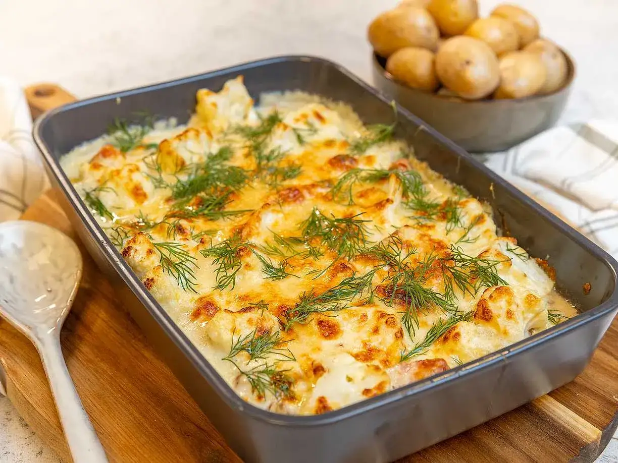 En ugnsform med kr&auml;mig stuvad blomk&aring;l, gratinerad med ost och toppad med dill. Bredvid syns en sk&aring;l med kokta potatisar.