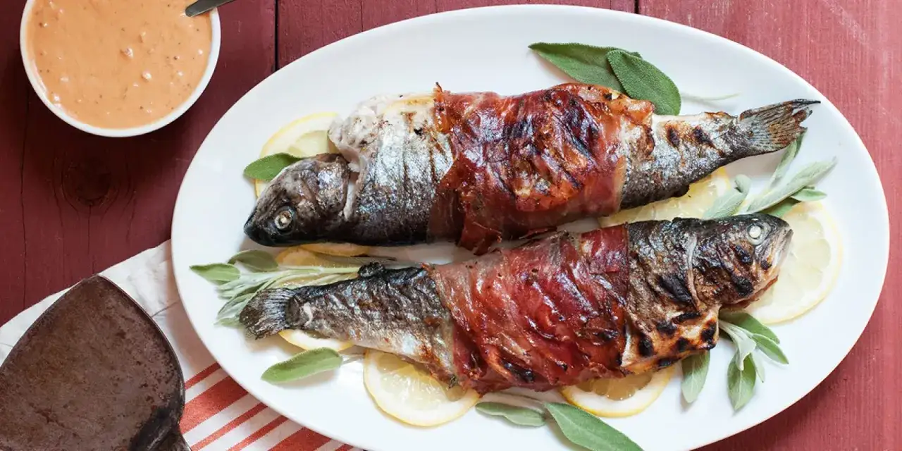 Zwei ganze Forellen, umwickelt mit Schinken, liegen auf einem Teller mit Zitronenscheiben und Salbeibl&auml;ttern. Ein Sch&auml;lchen mit Sauce steht daneben. Was ist crudo? Hier ist eine k&ouml;stliche Zubereitung!