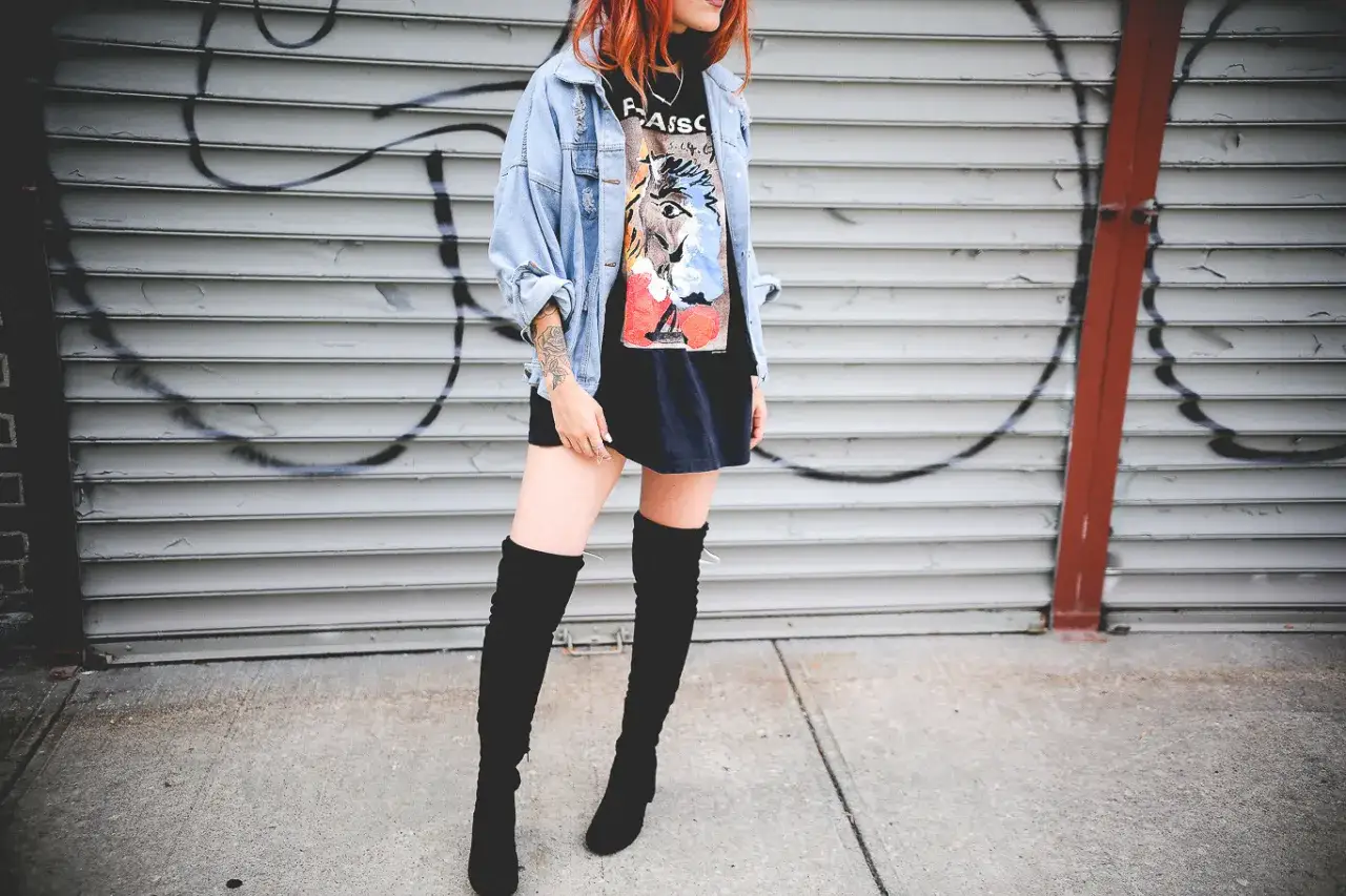 Chica con botas altas para bajitas, chaqueta vaquera y camiseta gr&aacute;fica, posando frente a una pared de garaje con grafitis.