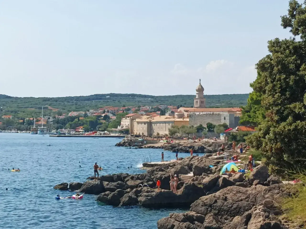 Słoneczna plaża na wyspie Krk, z kamienistym wybrzeżem, turkusową wodą i miasteczkiem w tle. Wiele osób korzysta z uroków wyspy Krk, podziwiając atrakcje.