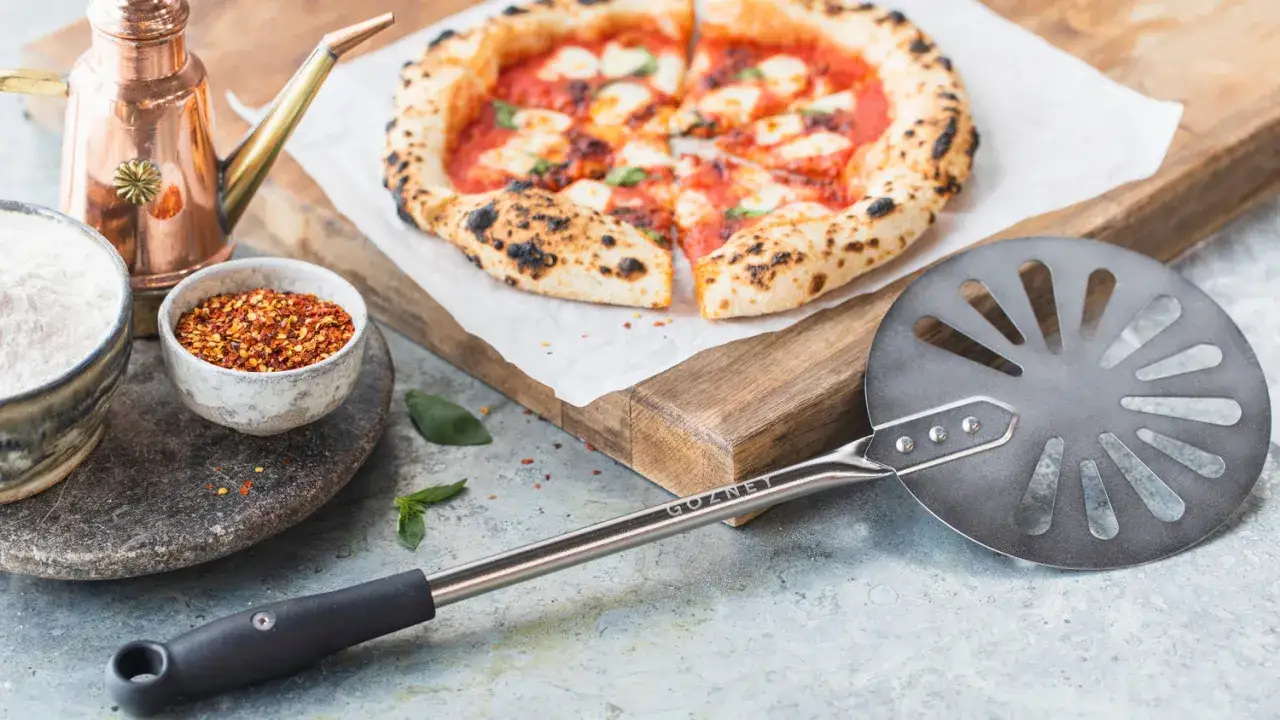 Perfekt pizzadeg till pizzasten. En nybakad pizza, mj&ouml;l, chiliflakes och en pizzaspade ligger redo f&ouml;r n&auml;sta m&auml;sterverk.