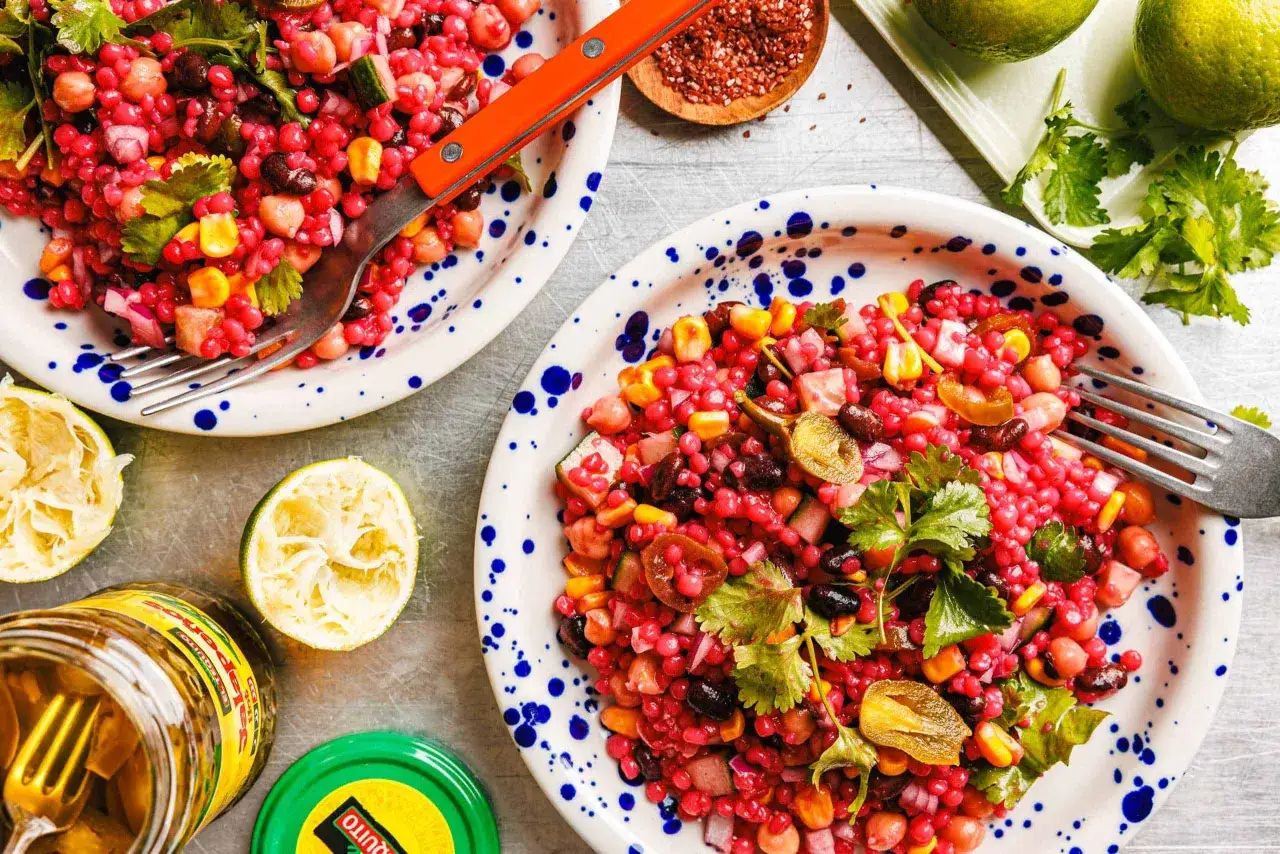 Bunter Couscous-Salat mit Kichererbsen, schwarzen Bohnen und Mais, garniert mit Koriander. Daneben liegen aufgeschnittene Limetten und ein Glas eingelegte Jalapeños.