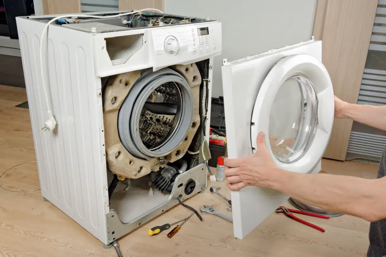 R&eacute;paration d'une machine &agrave; laver : une porte est retir&eacute;e, r&eacute;v&eacute;lant le tambour et les composants internes. Outils et cordon d'alimentation sont visibles.