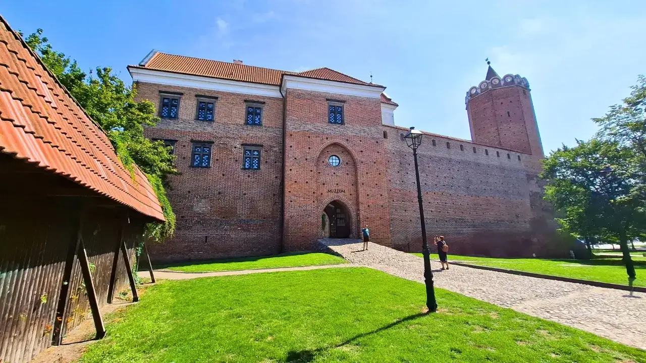 Zamek w Tumie, ceglana twierdza z wieżą, zaprasza do muzeum. Wokół zielony trawnik i brukowana ścieżka.