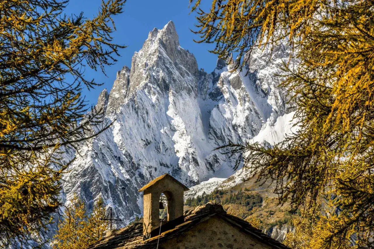 Piccolo campanile in pietra con campana, incorniciato da rami dorati di larice, con imponenti vette innevate del Val Ferret sullo sfondo.