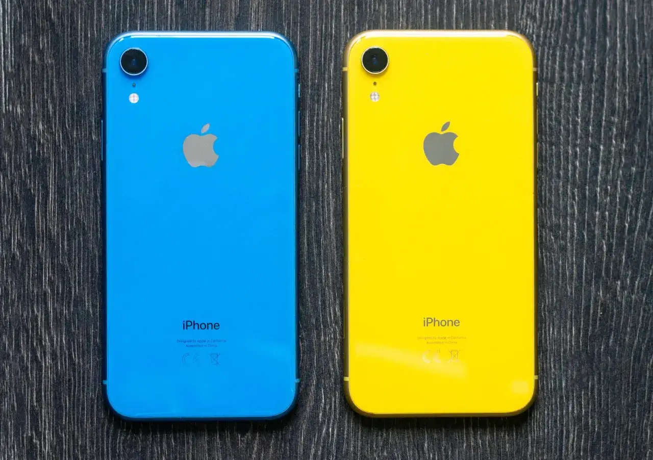 Dwa telefony iPhone XR z 2018 roku: jeden niebieski, drugi żółty, leżą na drewnianym tle.
