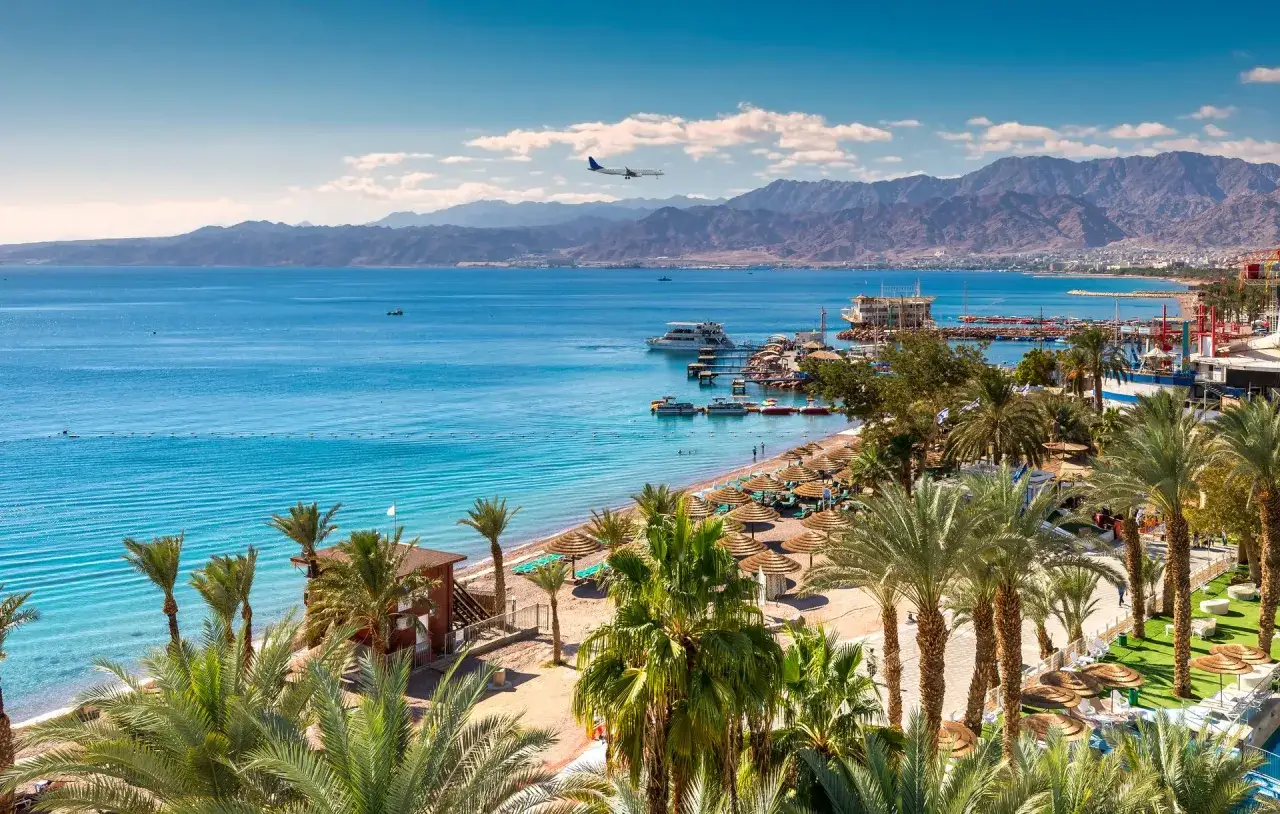 Piękna plaża z palmami i turkusową wodą Morza Czerwonego. W oddali góry, a nad nimi leci samolot. To widok na Eilat, gdzie leży Izrael.