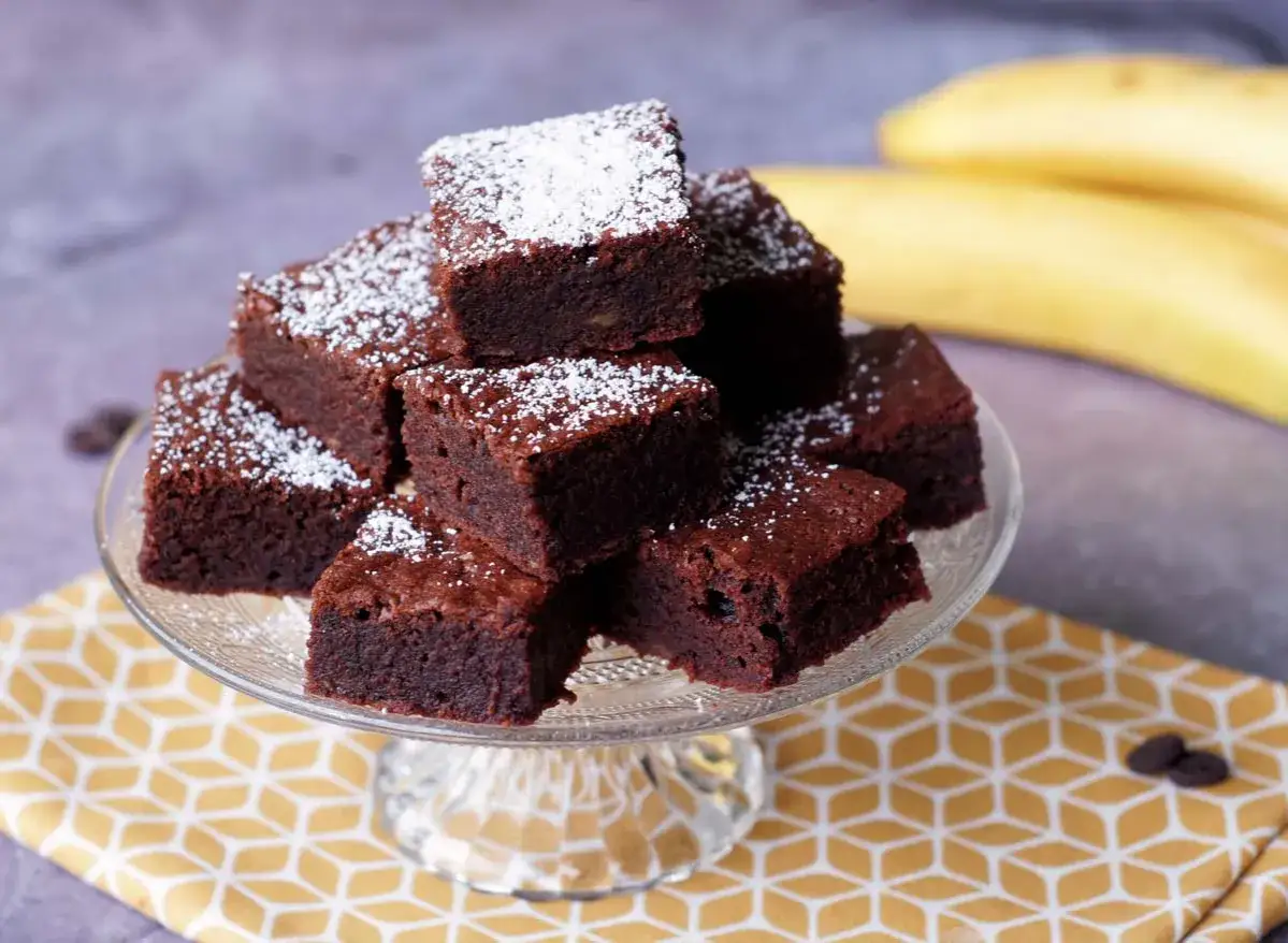 Saftige Bananen Brownies, best&auml;ubt mit Puderzucker, auf einem Glasst&auml;nder. Im Hintergrund reife Bananen.