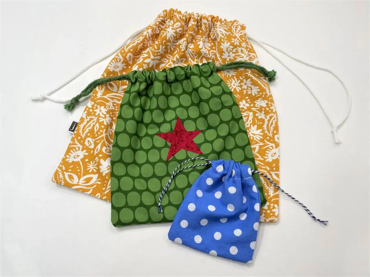 Drei bunte Beutel mit Kordelzug, inspiriert von einem Beutel Schnittmuster: gelb-weiß gemustert, grün mit roten Stern und blau mit weißen Punkten.