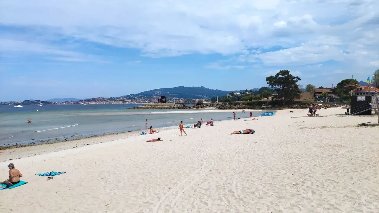 Gente disfrutando del sol y el mar en la playa Santa Marta de Baiona. El agua está tranquila y el cielo azul con nubes.
