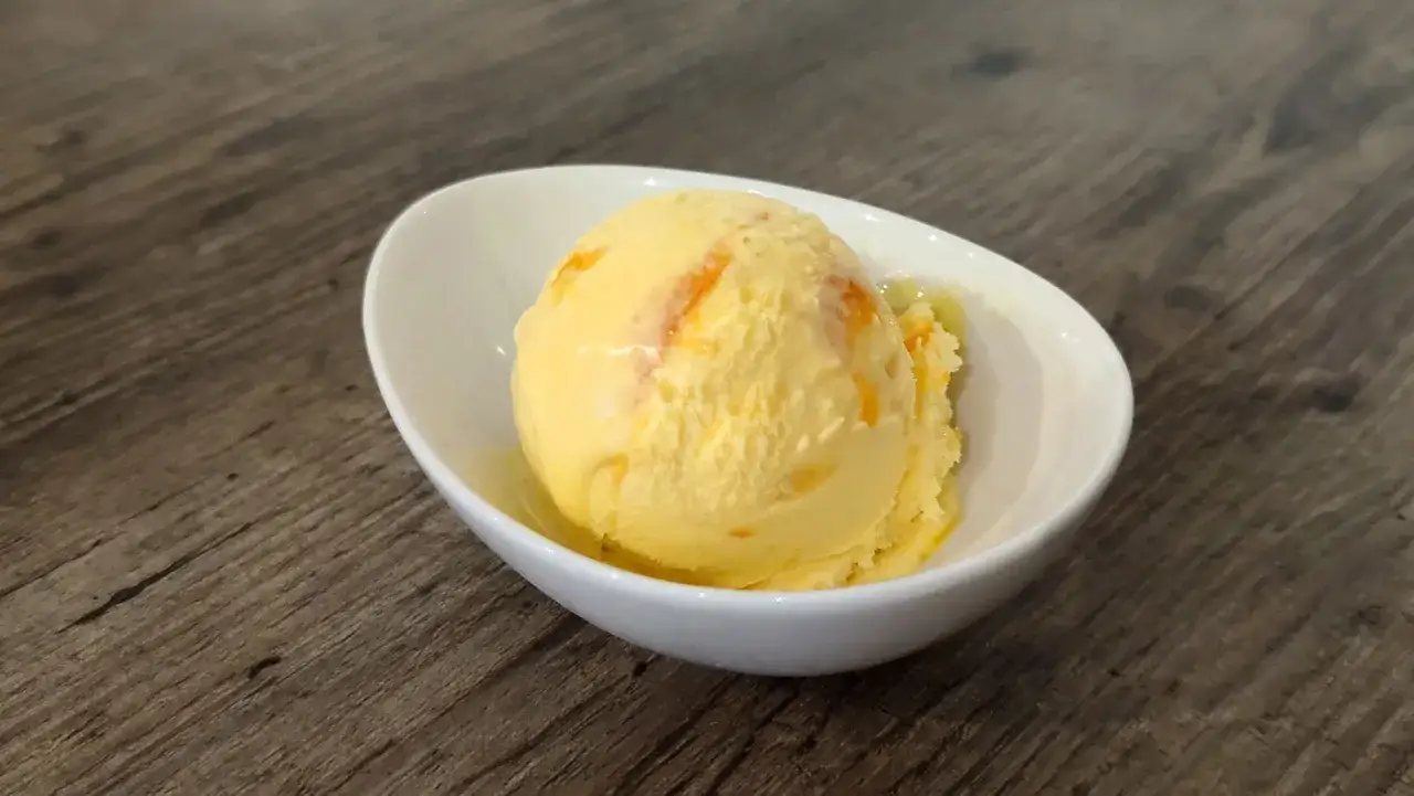 Ein Löffel cremiges Mandarinen Eis mit orangen Streifen in einer weißen Schale auf Holzhintergrund.
