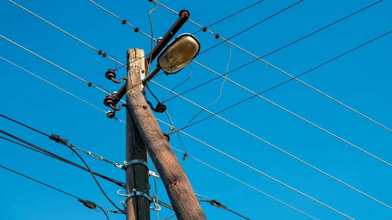 Drewniany słup energetyczny na działce z lampą uliczną i licznymi przewodami na tle błękitnego nieba.