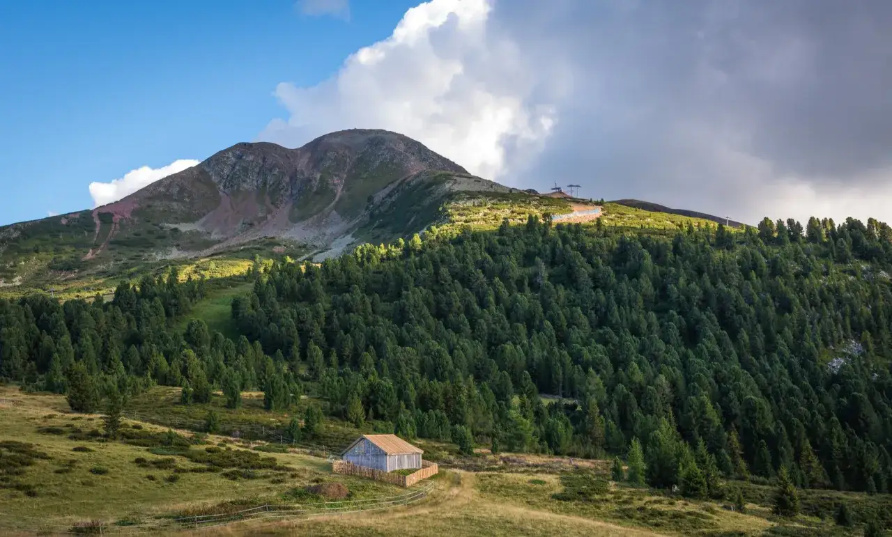Un rifugio solitario ai piedi del **Passo di Lavaz&egrave;**, circondato da un fitto bosco di conifere e montagne maestose sotto un cielo nuvoloso.