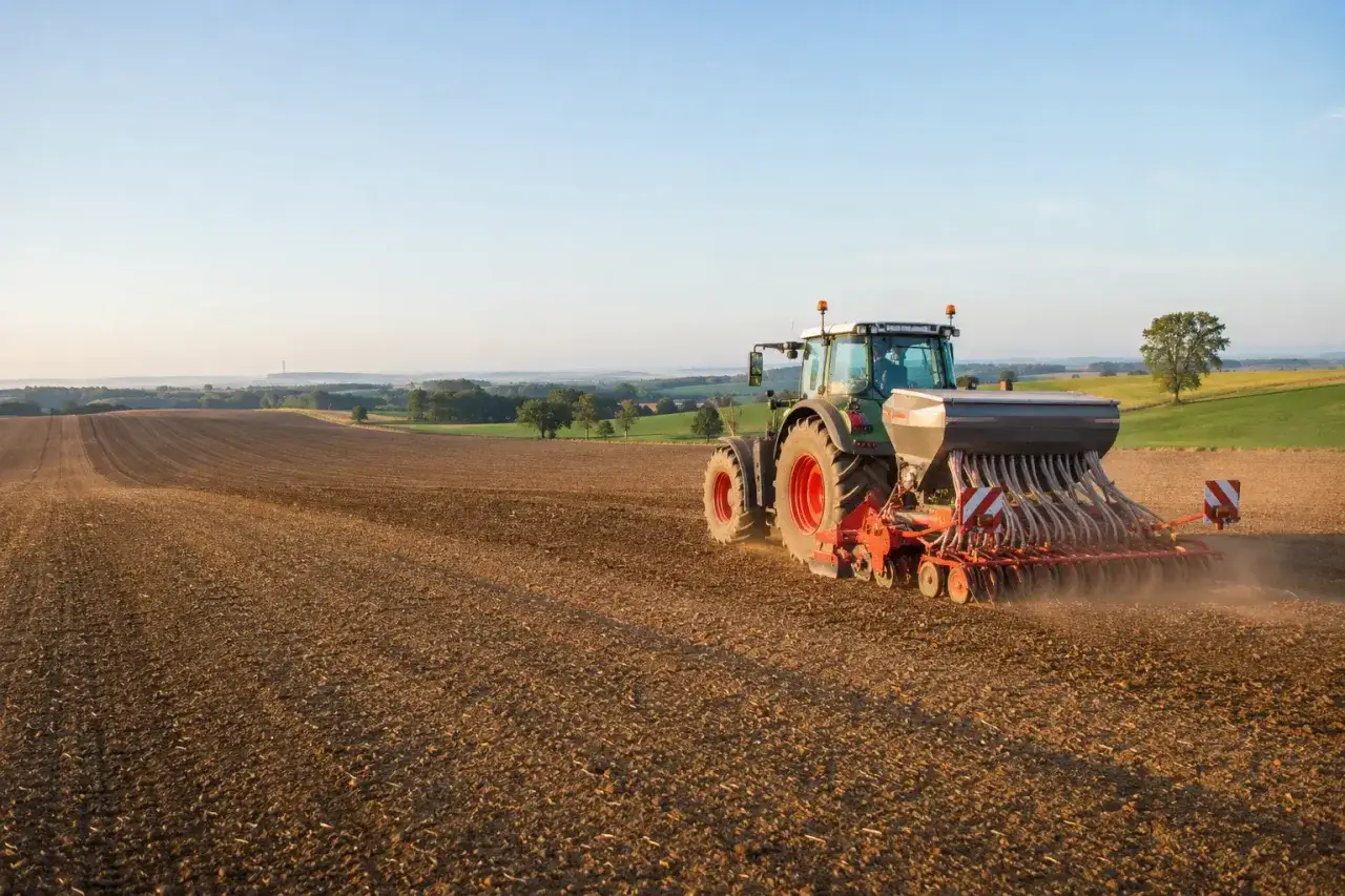 Traktor z siewnikiem pracuje na polu, siejąc rzepak. W tle łagodne wzgórza i błękitne niebo.