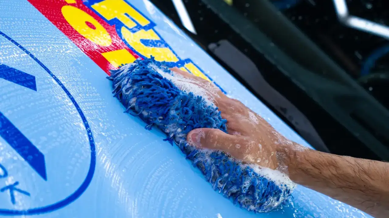 Mano che lava un'auto a casa con una spugna blu e schiuma. La carrozzeria è azzurra con scritte rosse e gialle.