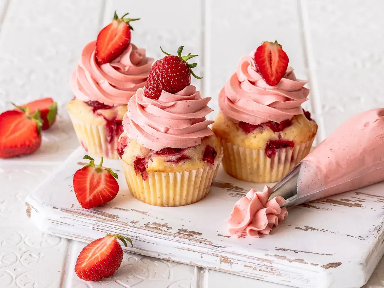 Drei Erdbeer-Cupcakes mit rosa Frosting und frischen Erdbeeren. Daneben eine Spritztüte mit Erdbeercreme und ein paar halbe Erdbeeren.