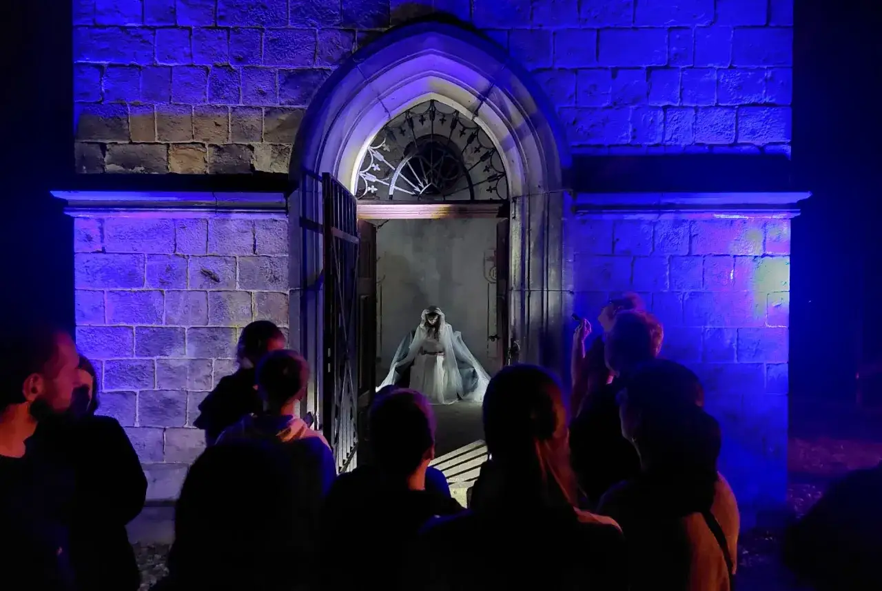 Widok na wejście do mrocznego budynku, gdzie postać w bieli czeka na widzów. Nocne atrakcje dla dorosłych.