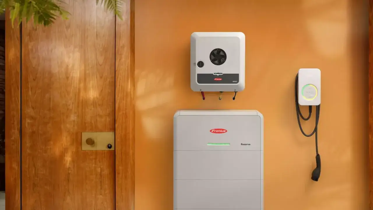 En Fronius v&auml;xelriktare och ett Fronius batteri monterade p&aring; en orange v&auml;gg. Bredvid syns en laddbox f&ouml;r elbil.